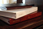 Three stacked hardcover books titled "THE RITUAL" on a dark wooden surface, with one white book in the middle and one dark red book at the bottom, under warm indoor lighting.
